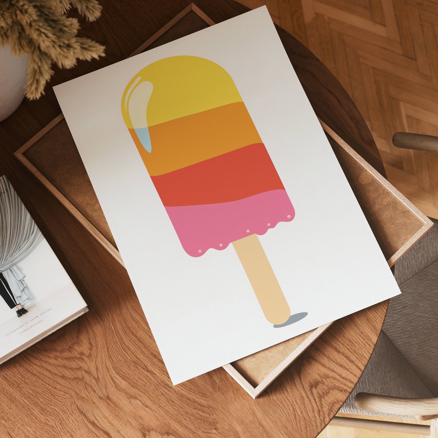 A printed poster, no-9, featuring a colorful popsicle with yellow, orange, red, and pink layers rests on a wooden table beside a book and a plant in a cozy, modern room.
